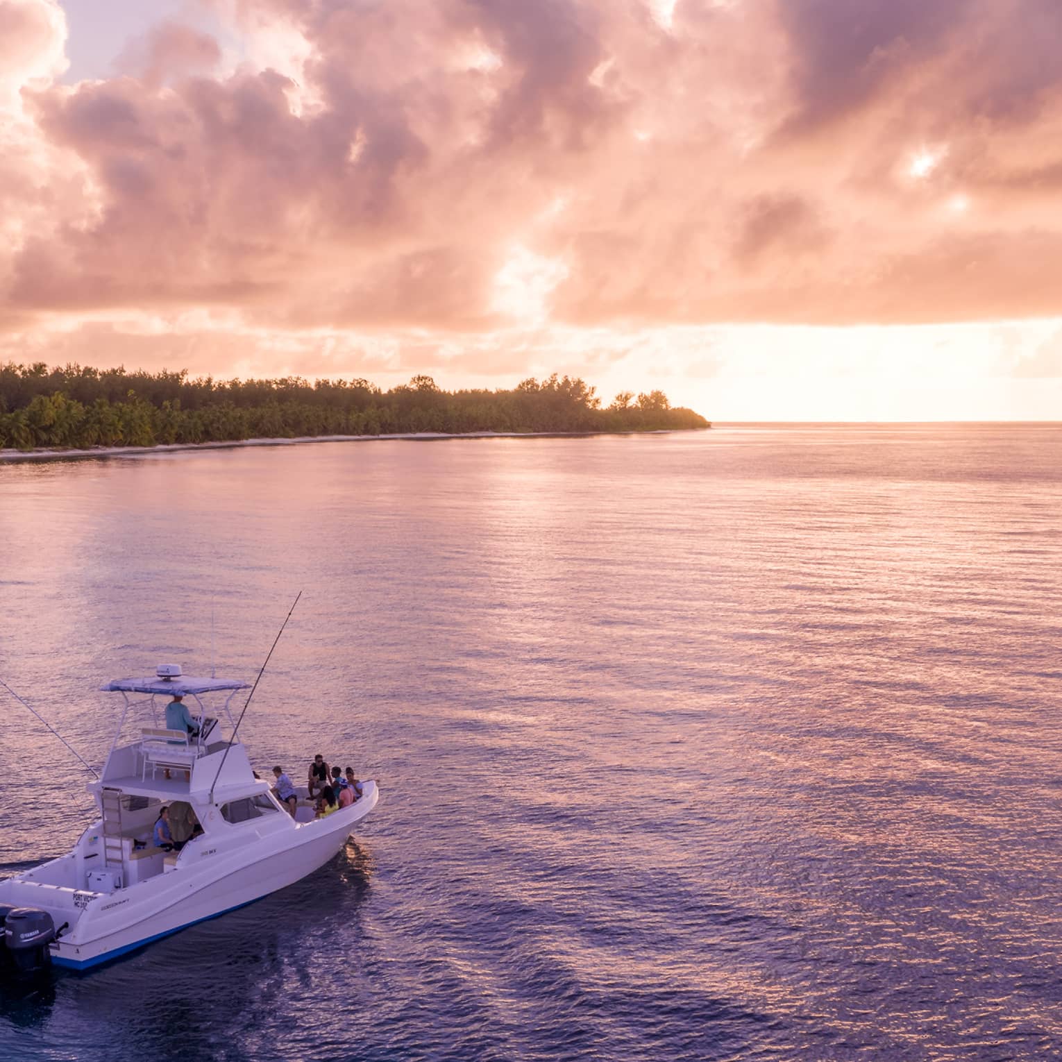 A group gathers at a boat's bow, drifting through calm water, a forested beach island and stunning sunset in the distance.