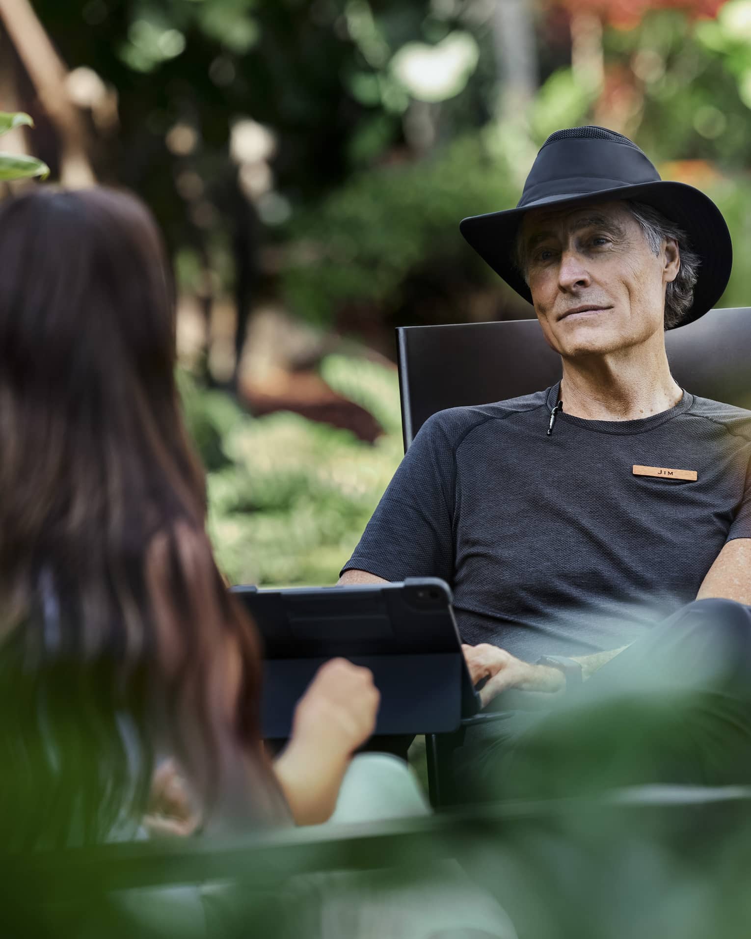 Sitting amid lush vegetation, two people engage in conversation, one with kind eyes and a soft smile typing on a tablet.