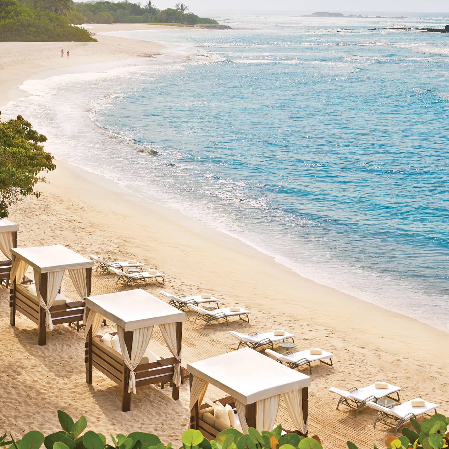 Amid lush vegetation, azure waves roll onto a curved beach lined with chairs and daybeds. A couple strolls in the distance.