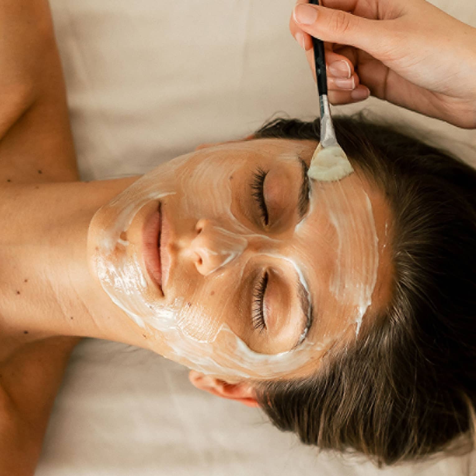 A person holding a small brush applies a white face mask treatment onto a woman's face