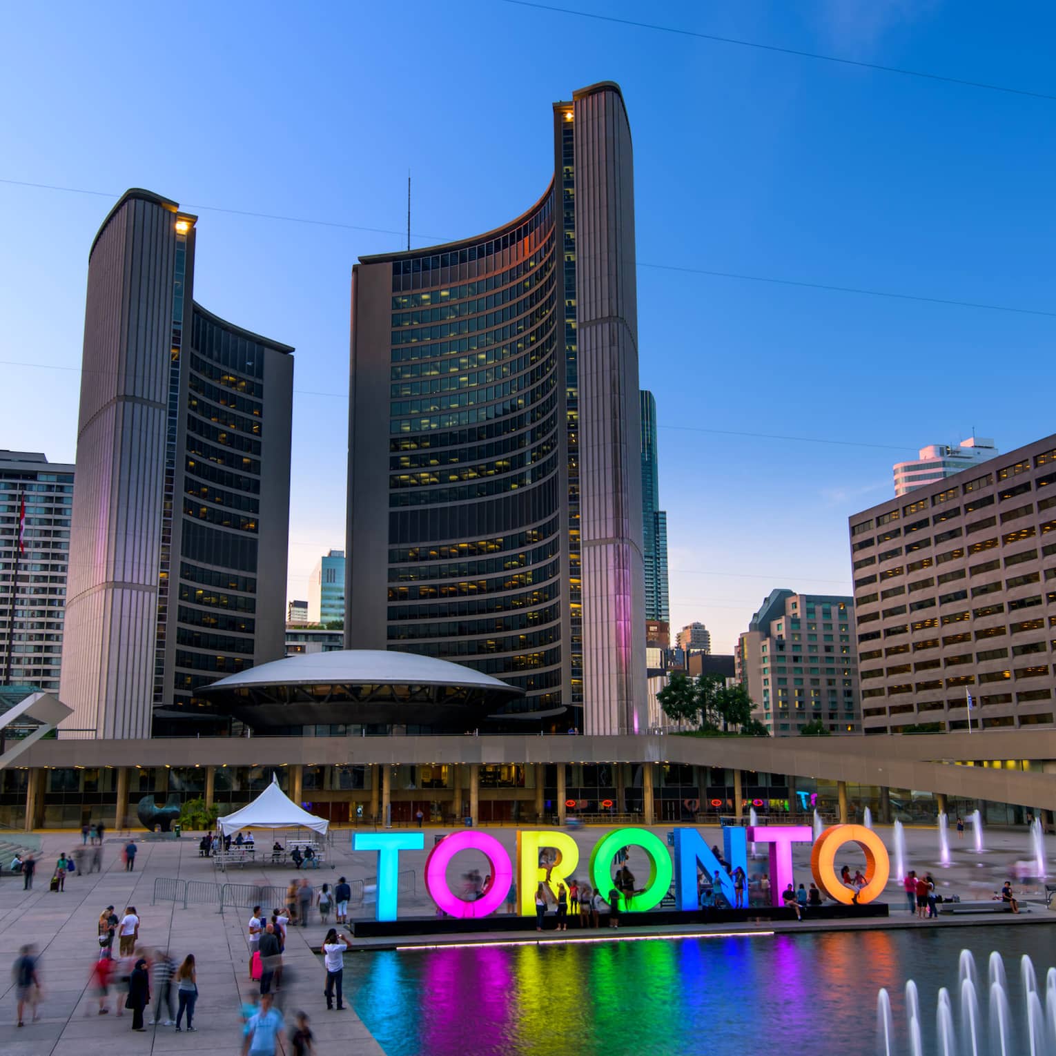 Colourful glowing letters spelling Toronto by fountain people in courtyard under curved City Hall buildings