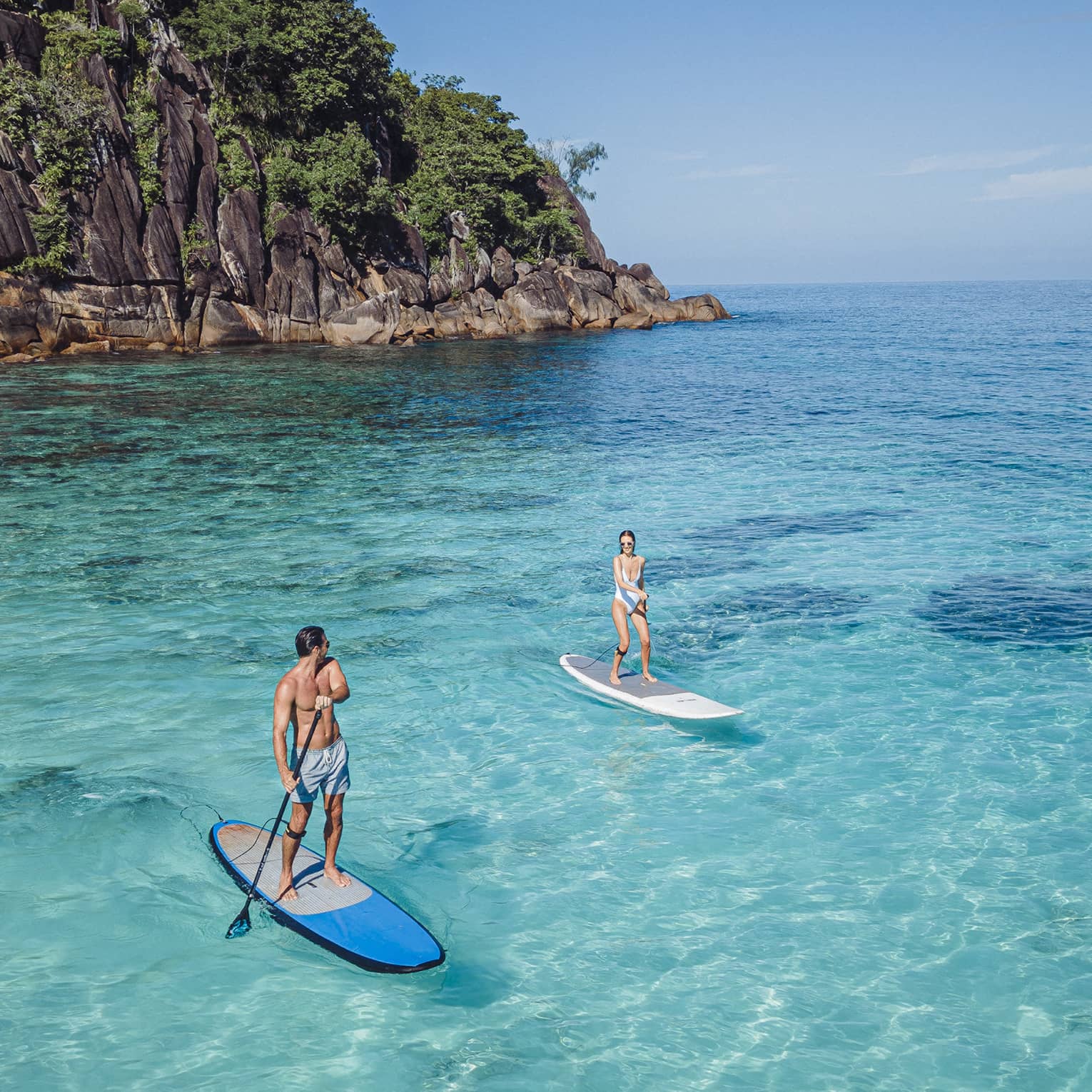 Two people stand-up paddleboarding on clear blue water with rocky cliff in back drop