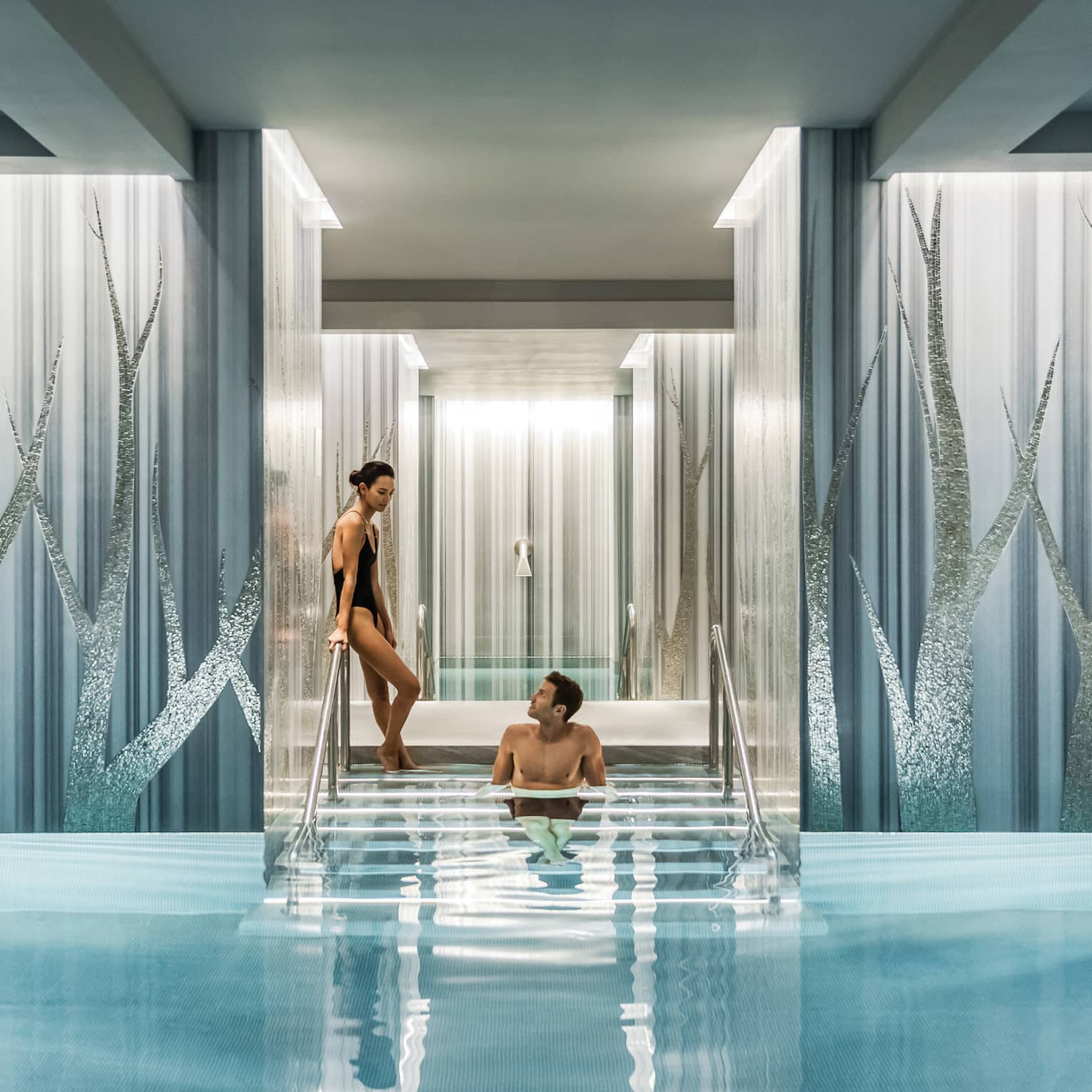 A couple lounges on the steps of a luxurious, serene indoor pool flanked by wide pillars decorated with a sparkly tree motif.