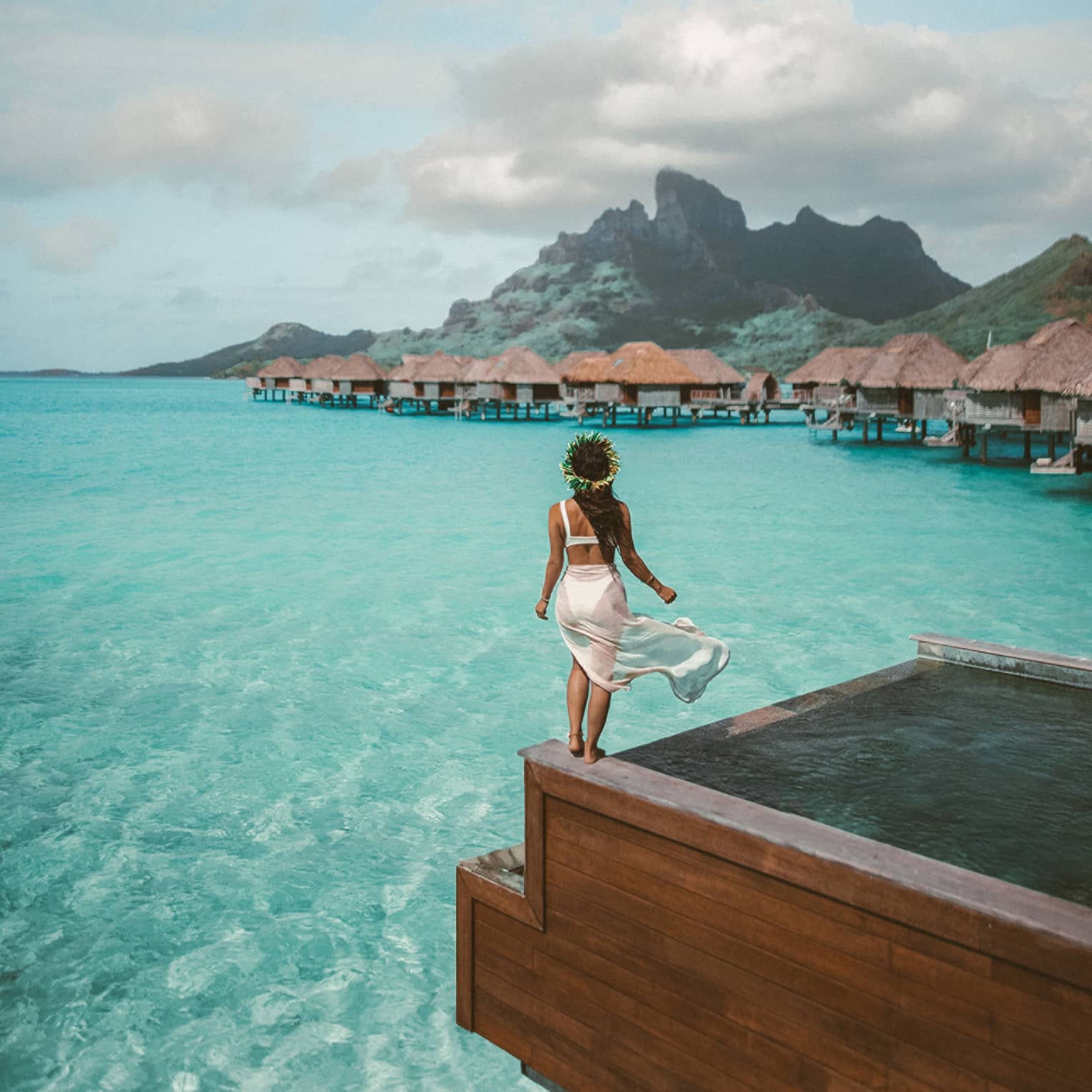 Woman stands at the edge of the infinity pool overlooking the overwater bungalows, mountains in distance