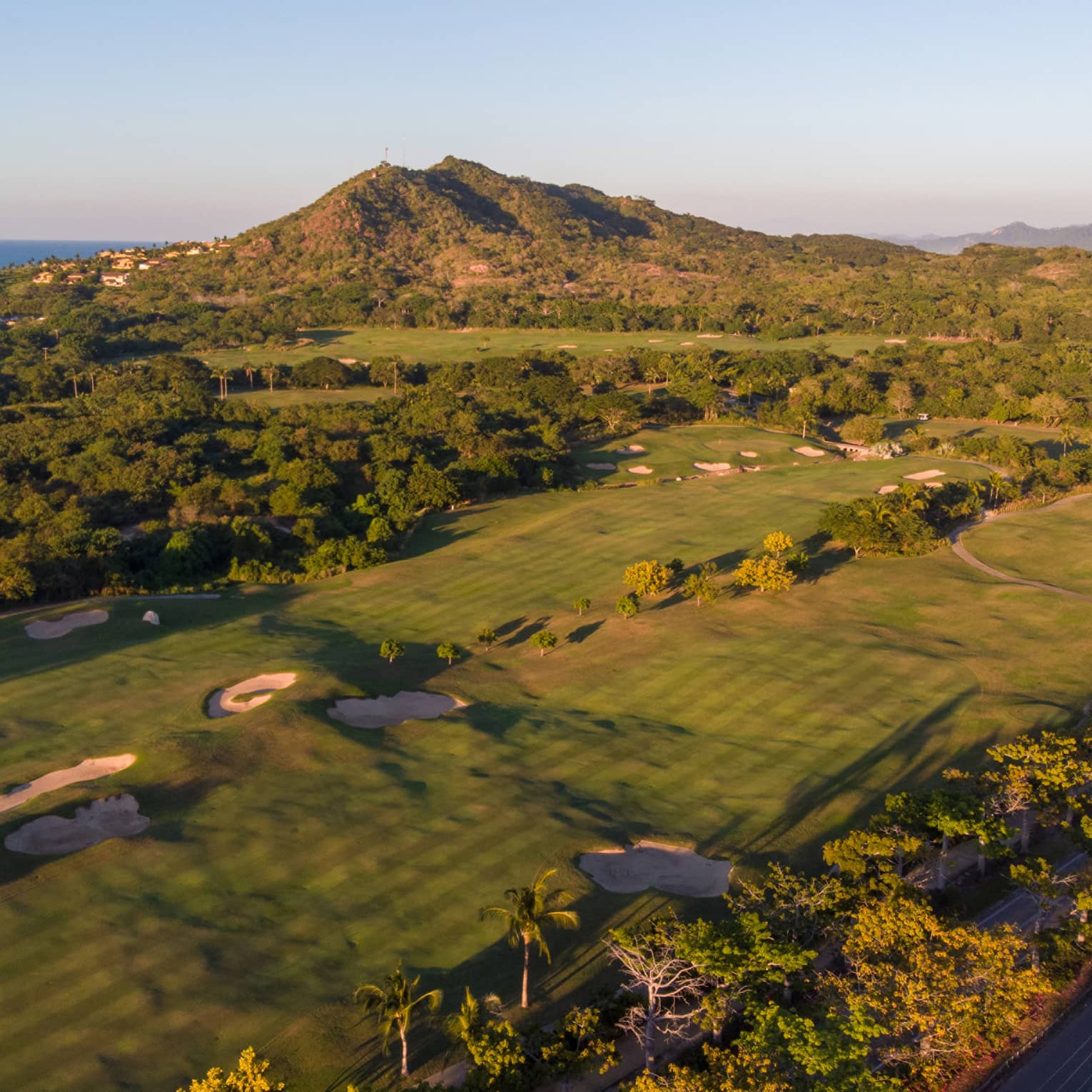 Aerial view of a golf course over the rolling coastal landscape; houses rising along the hillside, ocean in the distance.