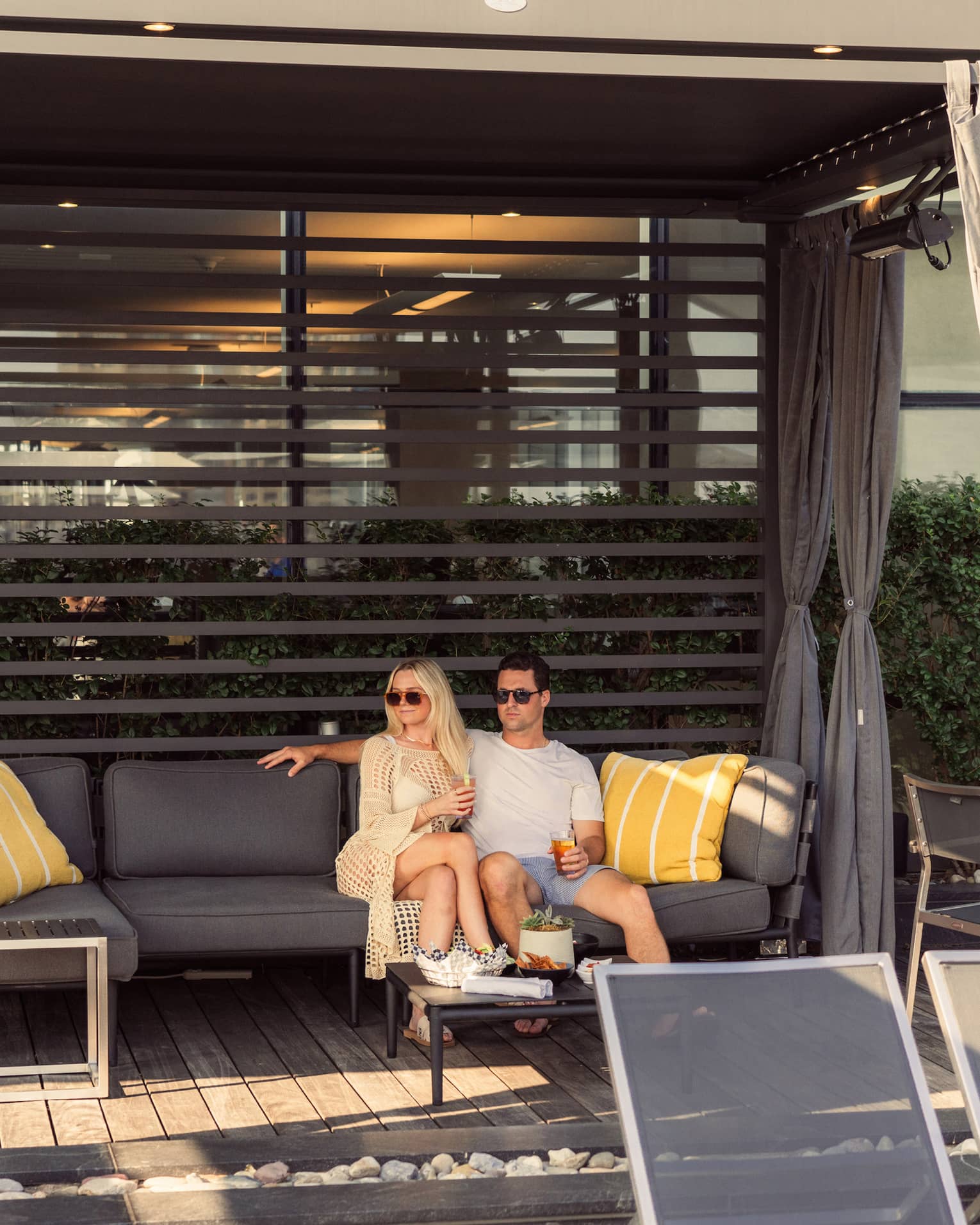 In a sun-dappled cabana, a couple relaxes together, drinks in hand, on a plush patio sofa, a table of snacks set for them.