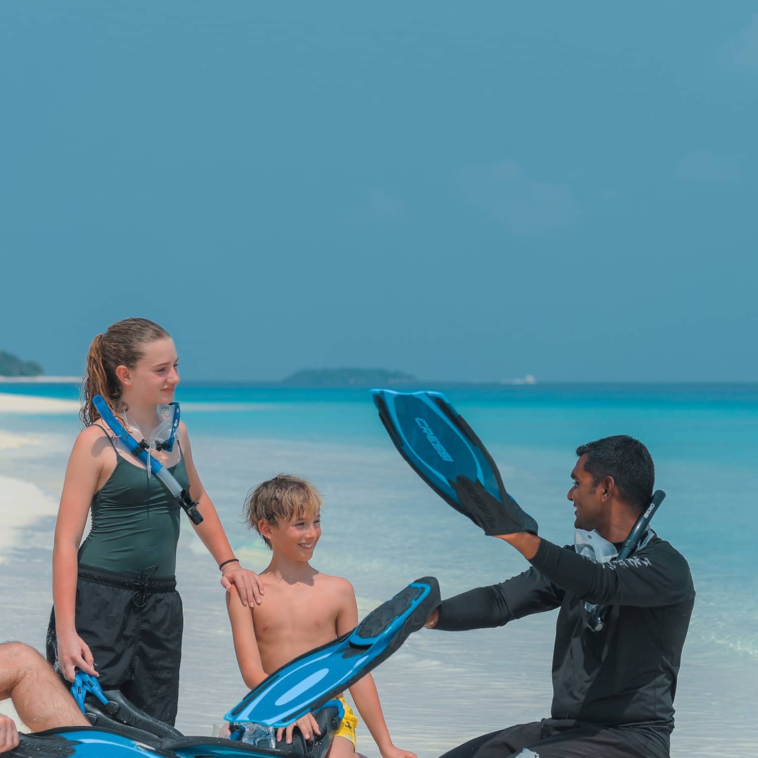 Sitting on a white sandy beach by the water, a family with snorkel gear intently listens to the instructor holding flippers.