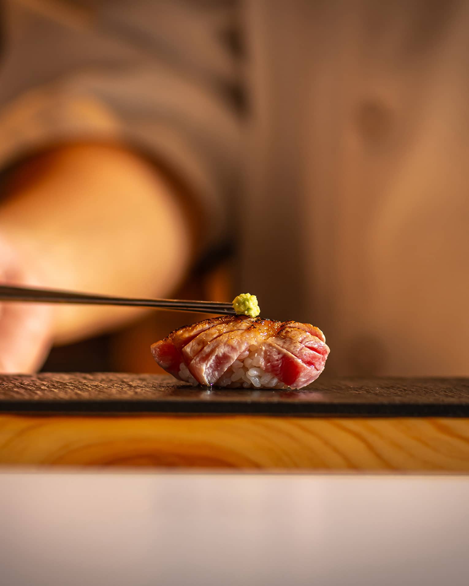 Seared tuna nigiri topped with a small dab of wasabi, placed on a black slate plate with a warm, blurred background