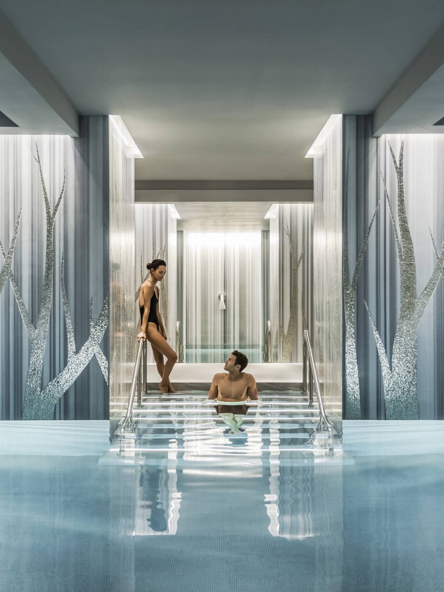 Woman in black swimsuit enters serenity pool while man sits on steps between blue and silver columns