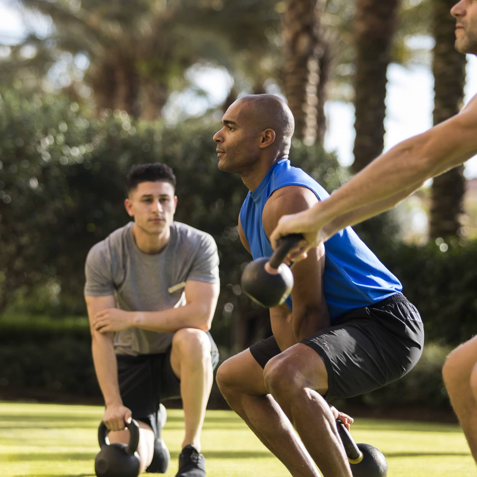 Four Seasons trainer watches as two men squat, lift kettlebell weights on lawn