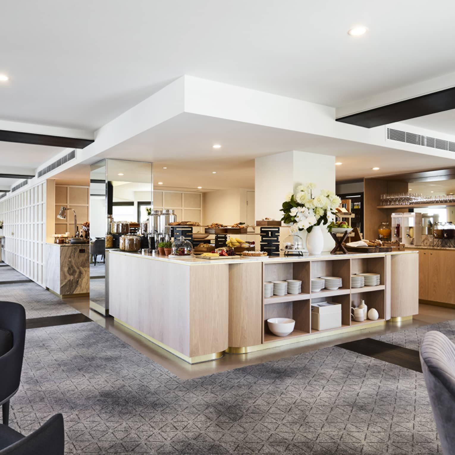 A modern hotel lounge with plush grey chairs and carpet, and a well-equipped kitchen set up with buffet foods and coffees.
