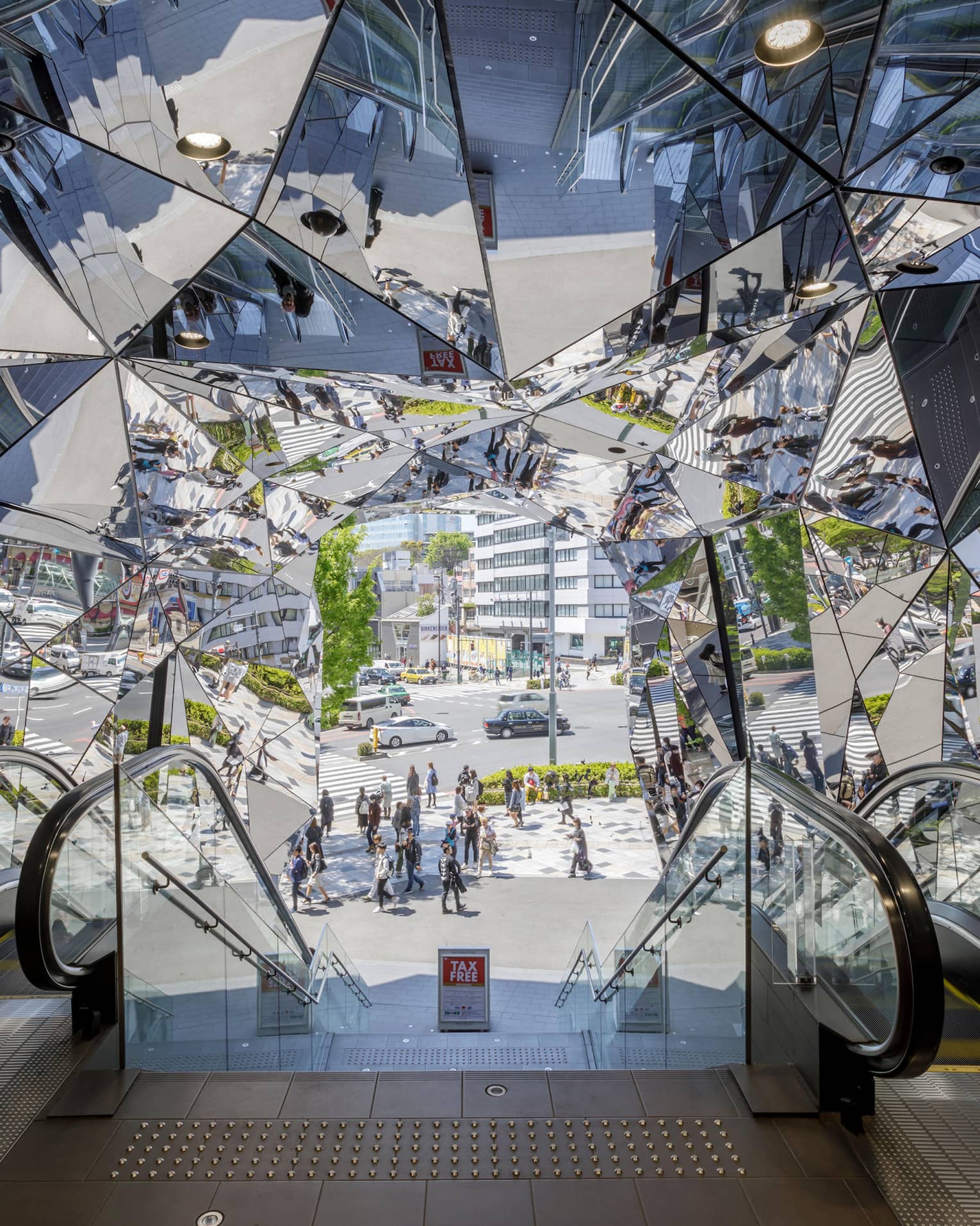 Escalators leading to a mirrored entrance reflecting pedestrians and a busy street scene
