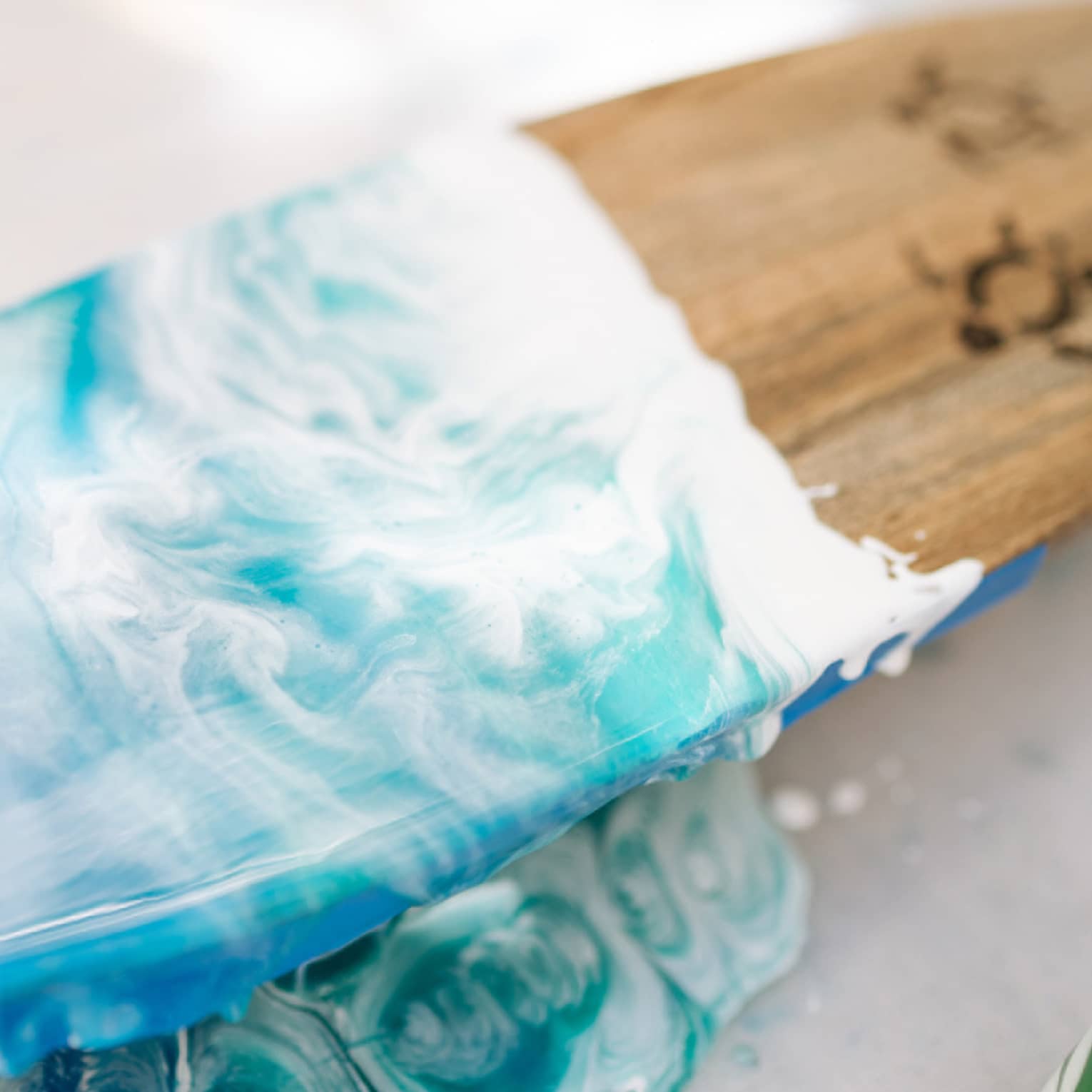 Close-up of wooden surfboard half-painted in deep to turquoise blues with white wave-like swirls and turtle outline drawings.