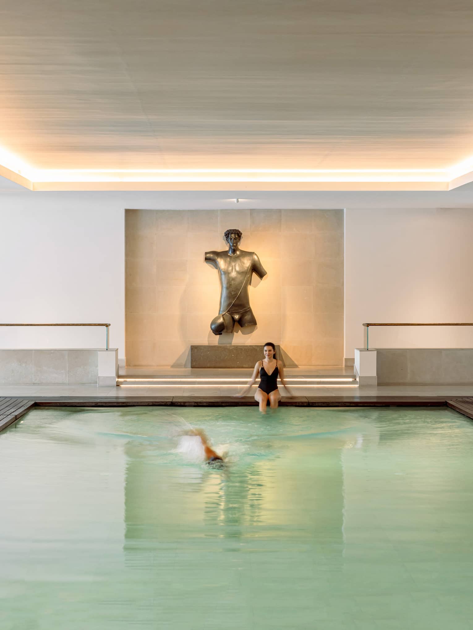 Person sitting at end of indoor lap pool under uplit ceiling while another person swims