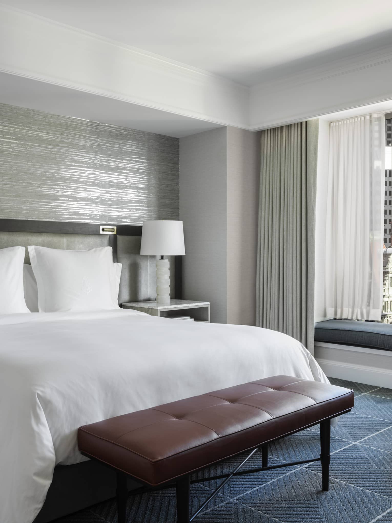A hotel room with a bed with white sheets, a red bench, a grey chair, two white lamps, and a large window looking out at a city.