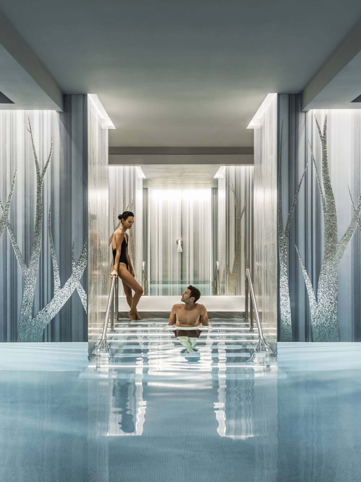 Woman in black swimsuit enters serenity pool while man sits on steps between blue and silver columns