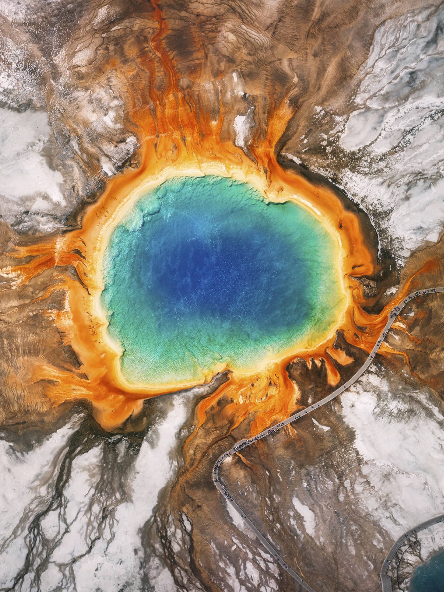 Aerial view of the Grand Prismatic Spring in Yellowstone, featuring vibrant blue, green, yellow and orange concentric rings, surrounded by rocky terrain