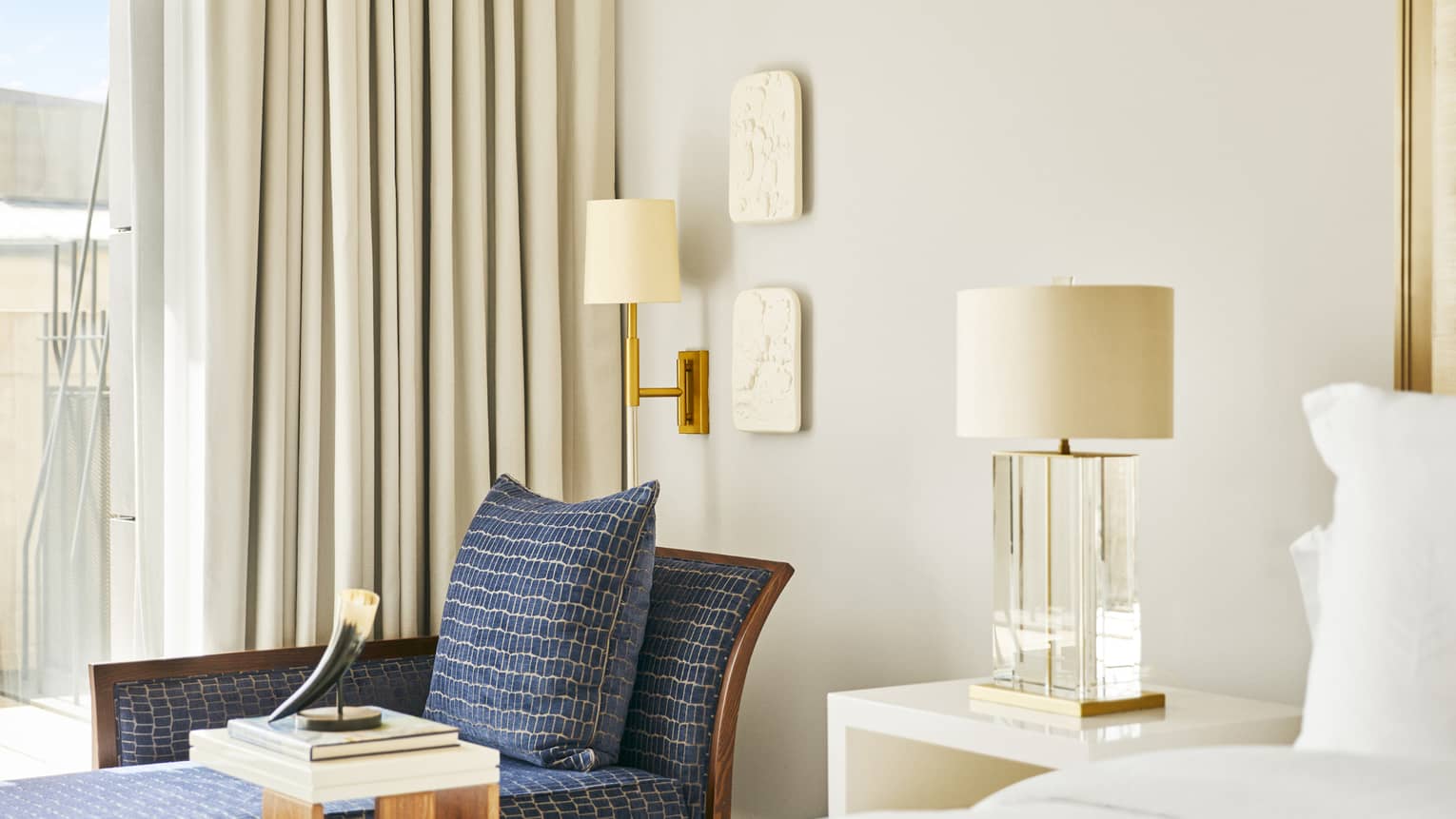Luxury hotel bedroom corner with blue chaise lounge, horn sculpture on side table, wall-mounted lamp and bedside table with glass lamp