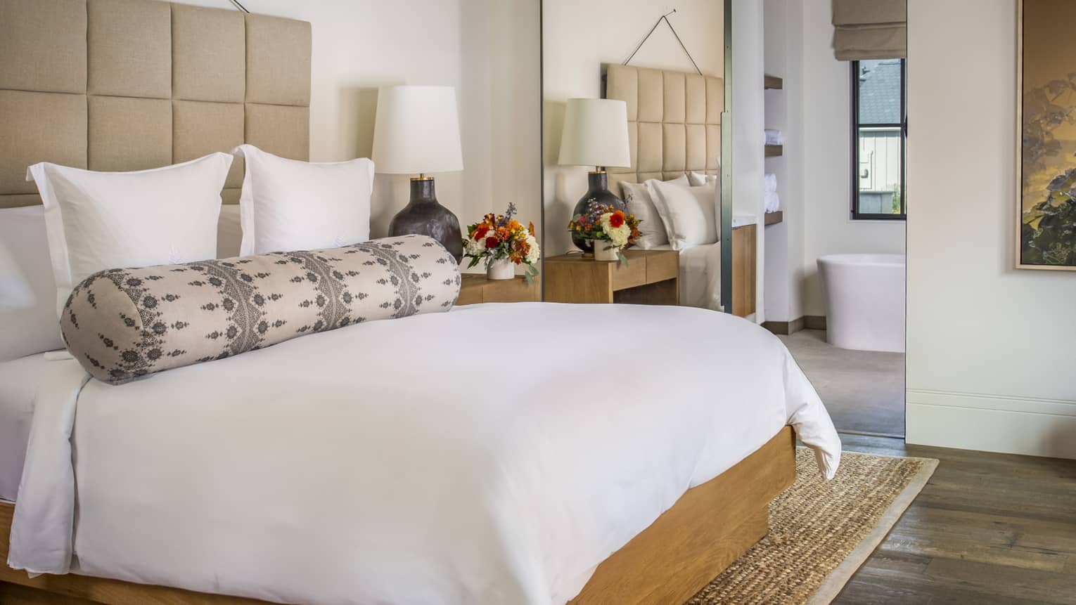 Resort bedroom with a king bed, beige upholstered headboard, patterned bolster pillow, wood nightstands with lamp and flowers and a view into bathroom with tub