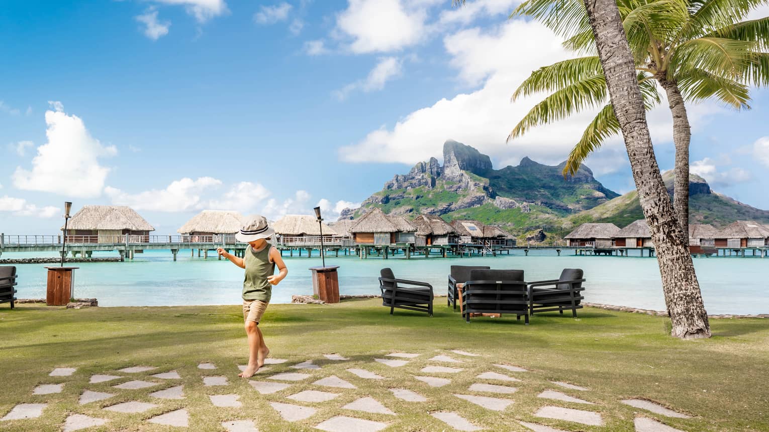 A child tiptoes on paving stones by a clear lagoon. Grass-roofed suites stand in the water in front of island mountains.