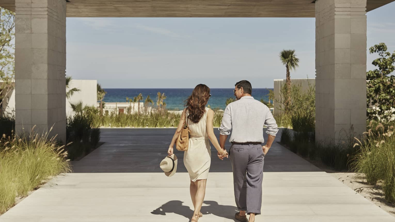 Rear view of a couple walking hand in hand towards a gradient blue ocean on a covered boardwalk lined by tall grasses.