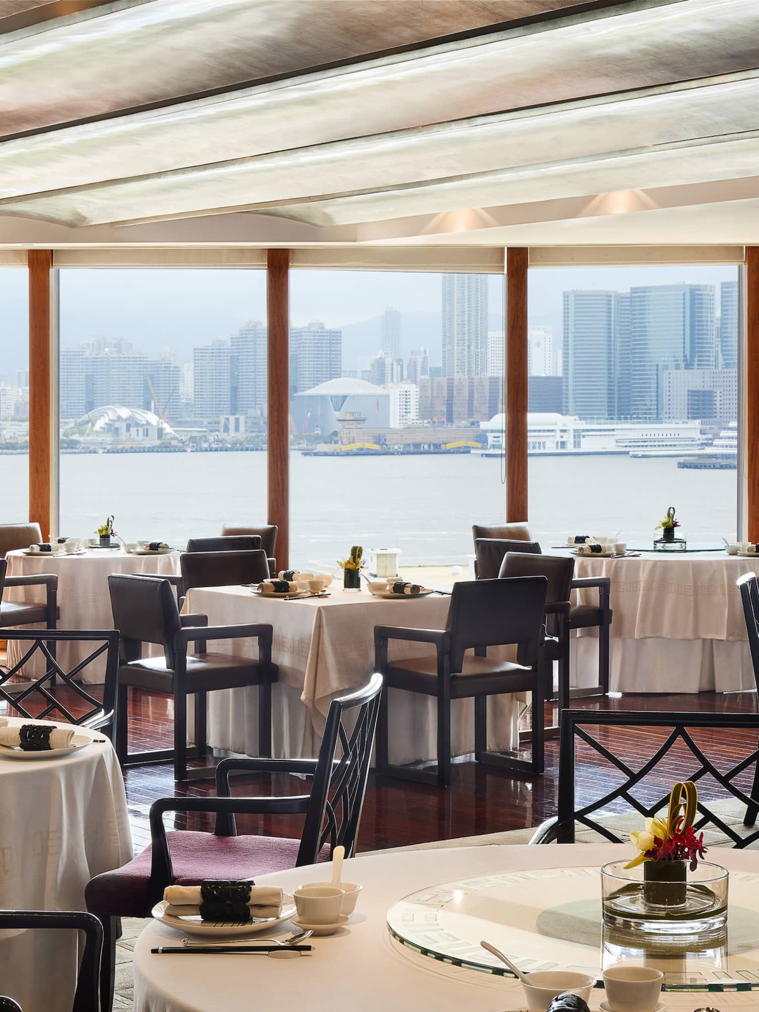 An elegant dining room with tables set with flowers and place settings, overlooking the city from across a body of water.