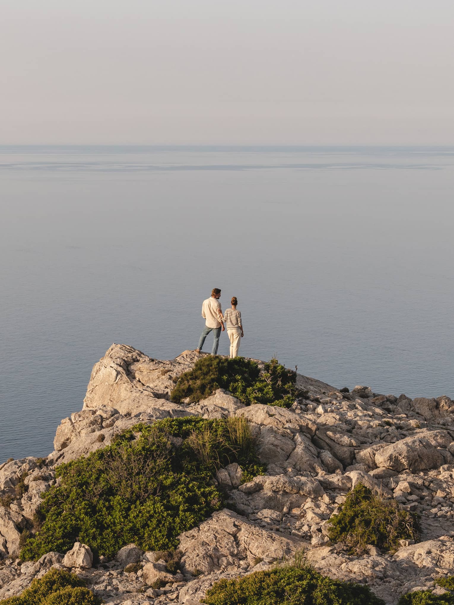 Distant view of a couple in hazy sunlight gazing at an expanse of tranquil water from atop a cliff dotted with shrubs.