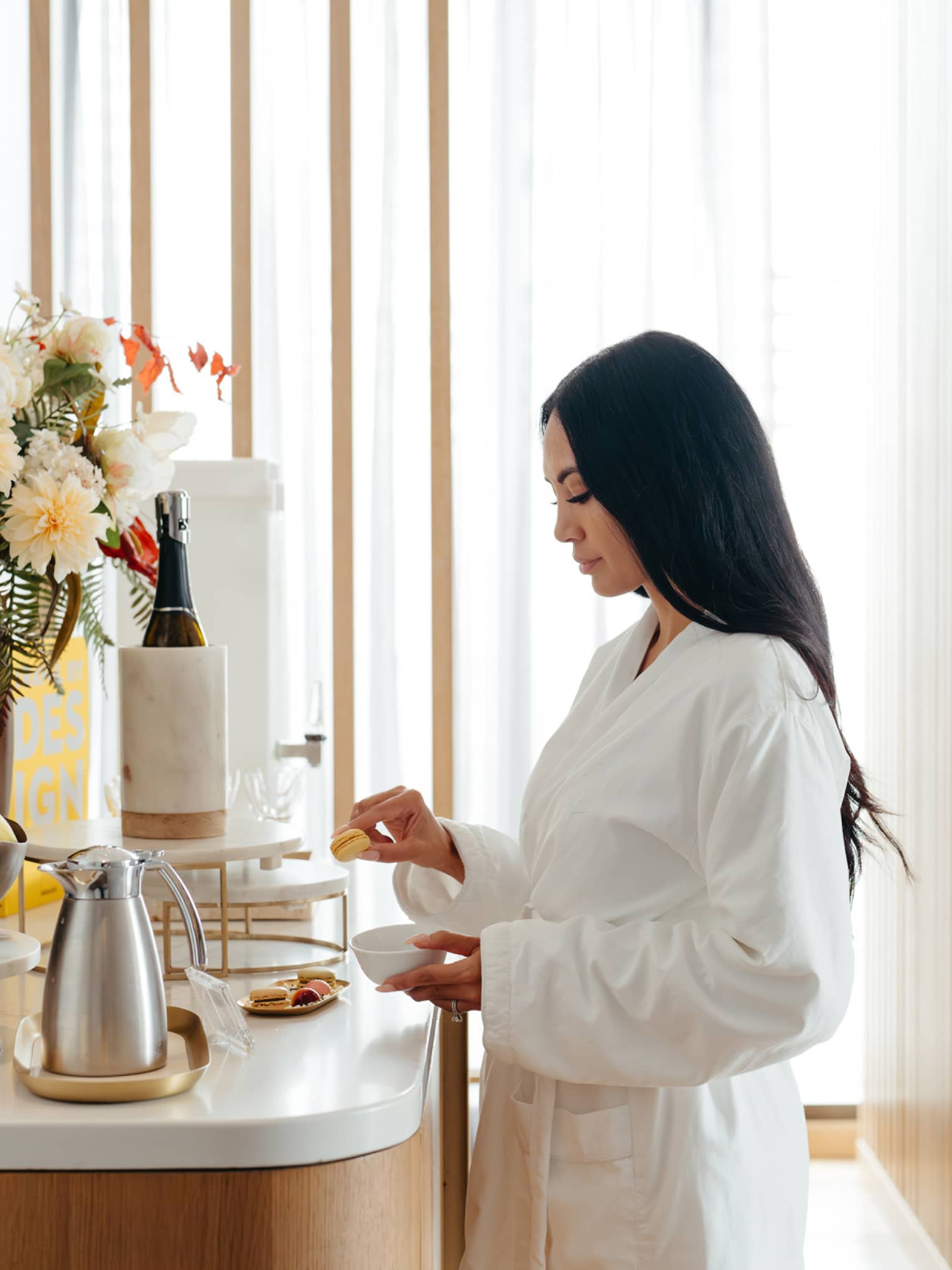 A guest in a white robe stands in a light-filled interior space in front of a counter holding a floral arrangement, a bowl of fresh fruit, a plate of small food items, a carafe and other beverage items.