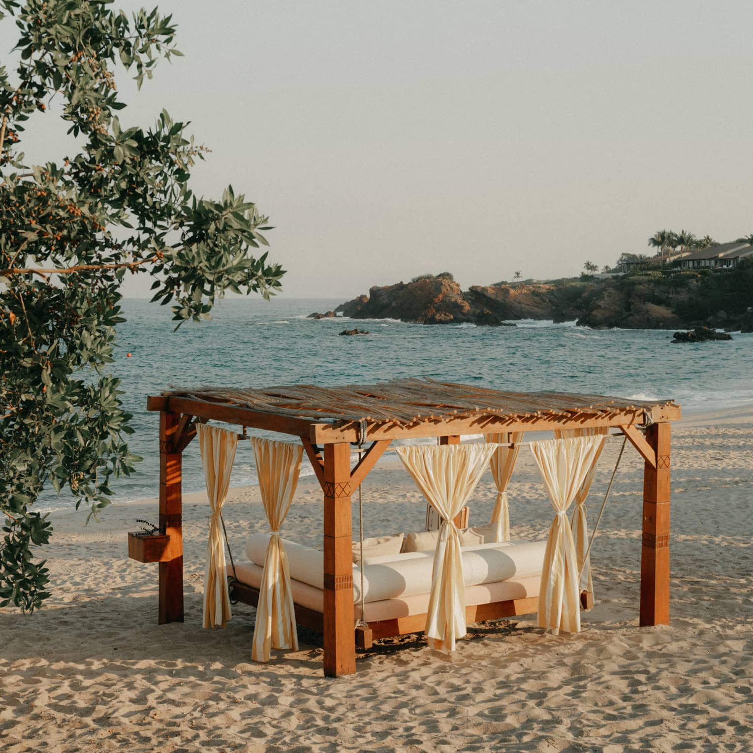 A cabana with curtains and a daybed, on a sandy beach next to stand of trees, with ocean and shoreline views in the background