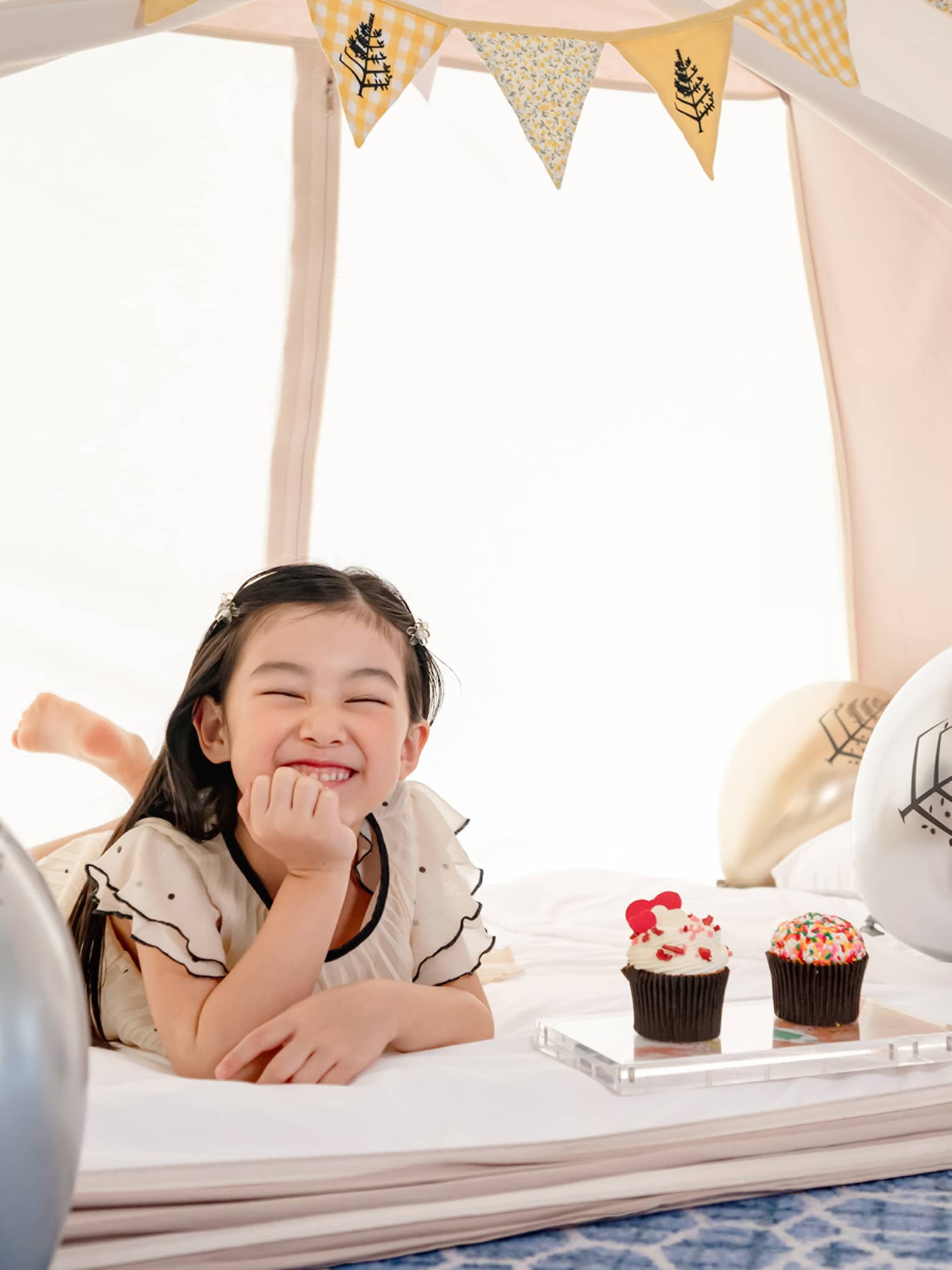 Smiling young girl in in-room kids? tent with gold and silver balloons and cupcakes