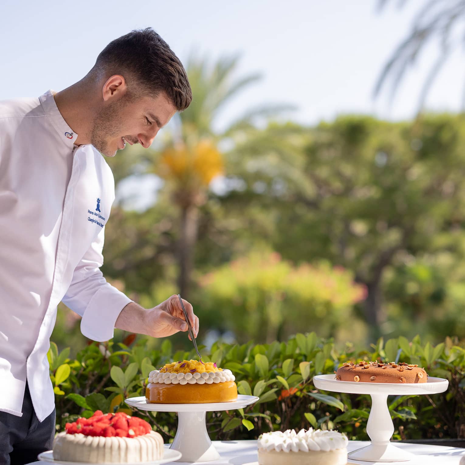 Chef in white chef's coat delicately placing garnish on cake atop cake stand among other desserts