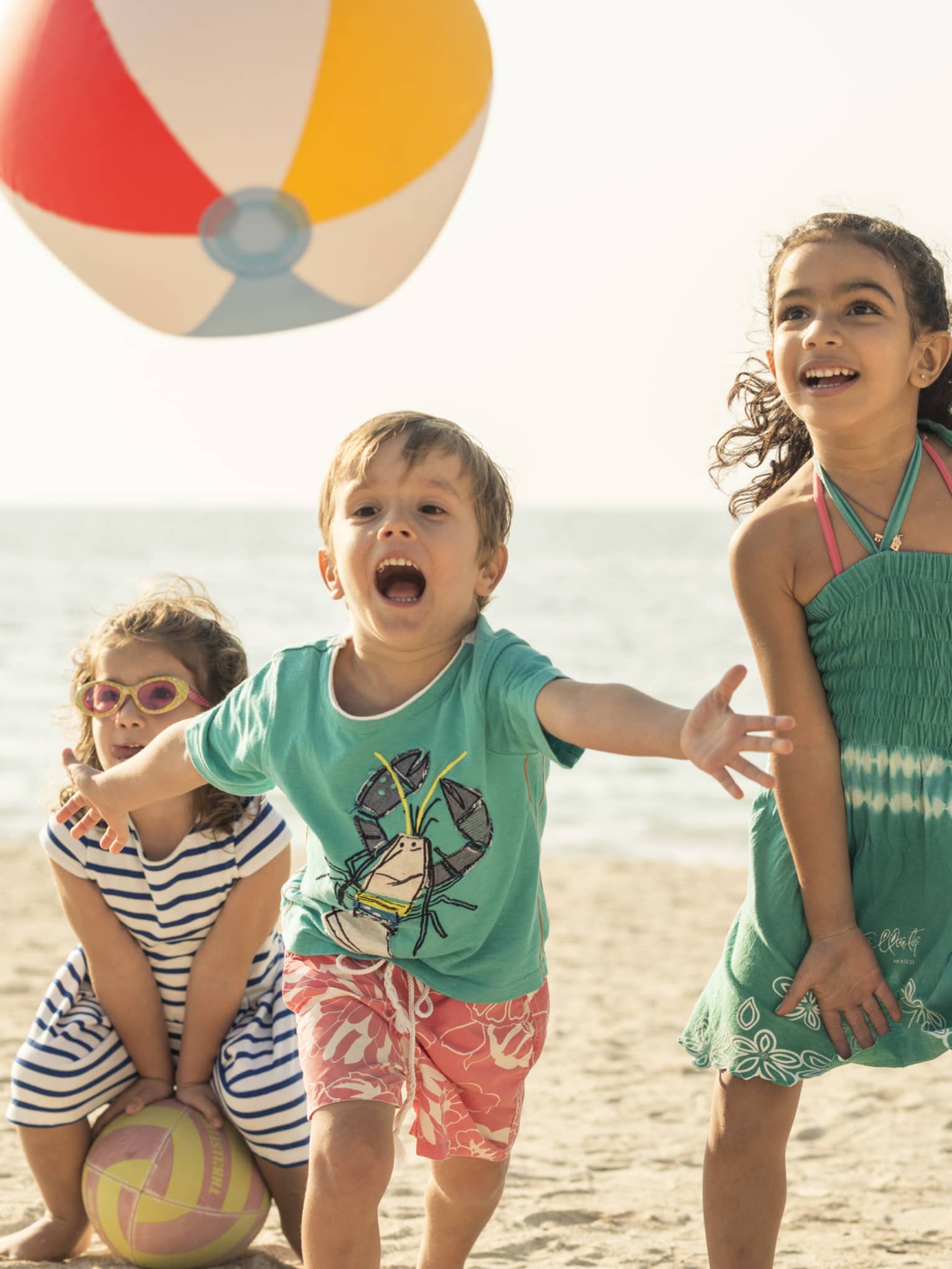 Close-up of three children playing with a beach ball on a sandy beach, the calm ocean and clear sky spanning behind them.
