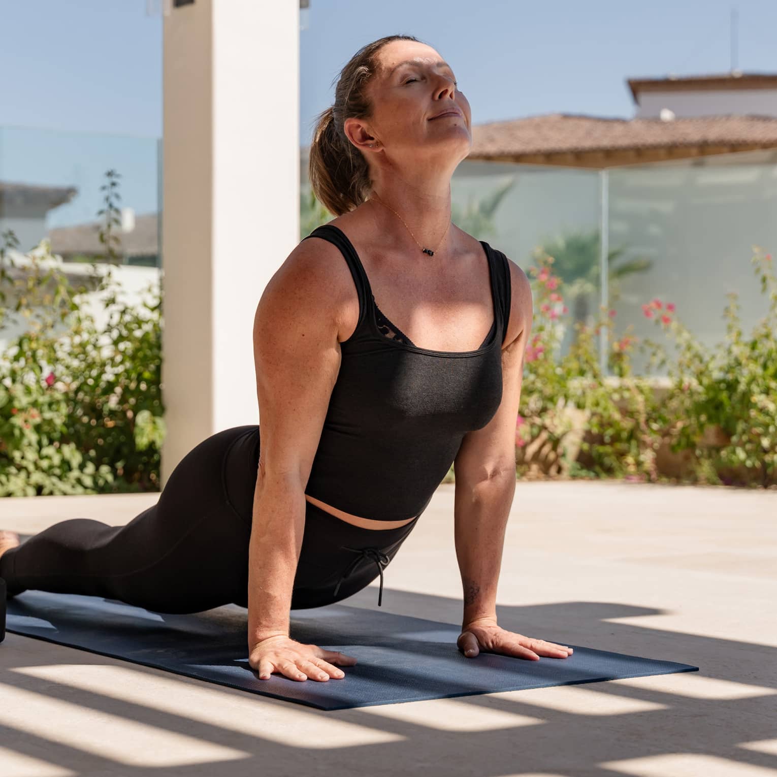 In the shade of a trellis, a guest gets a deep low back bend supported by their hands in an upward-facing-dog yoga pose.