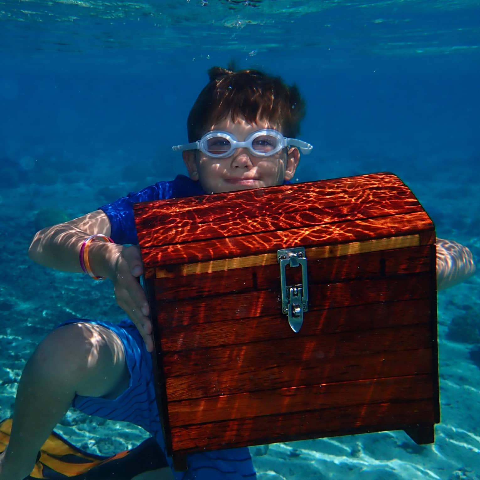 A underwater view of a child resting on the ocean floor, dressed in goggles and flippers and holding a treasure chest aloft.