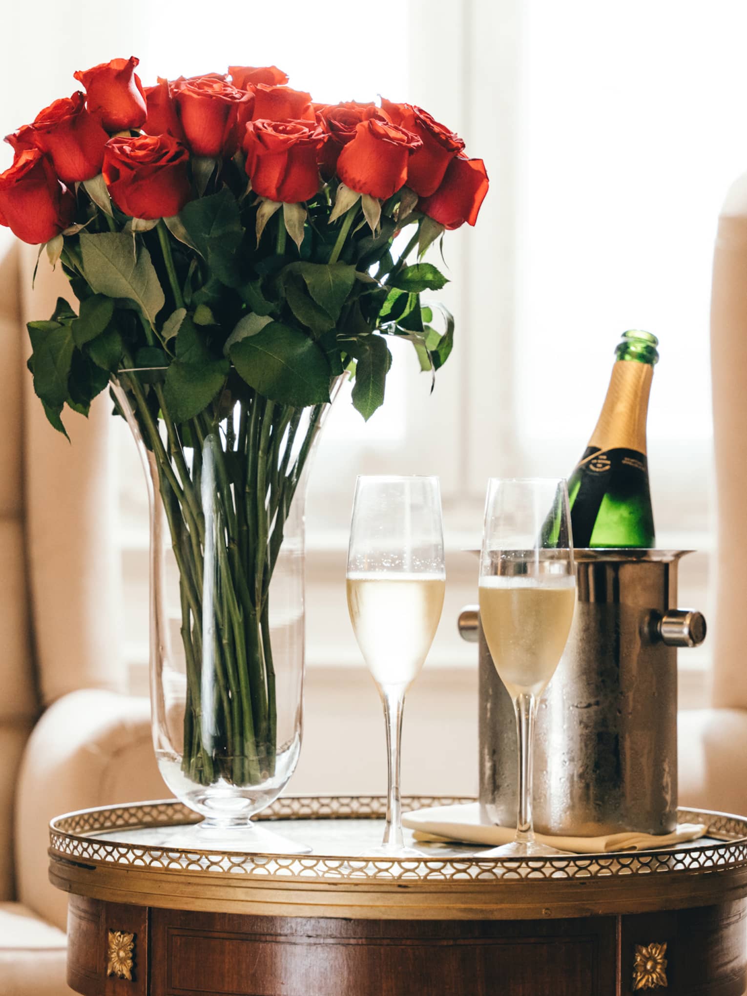 A glass vase filled with a bouquet of red roses, two flutes of champage and a champage botte in a silver holder sit on a small, round, gold table in front of two beige trufted arm chairs
