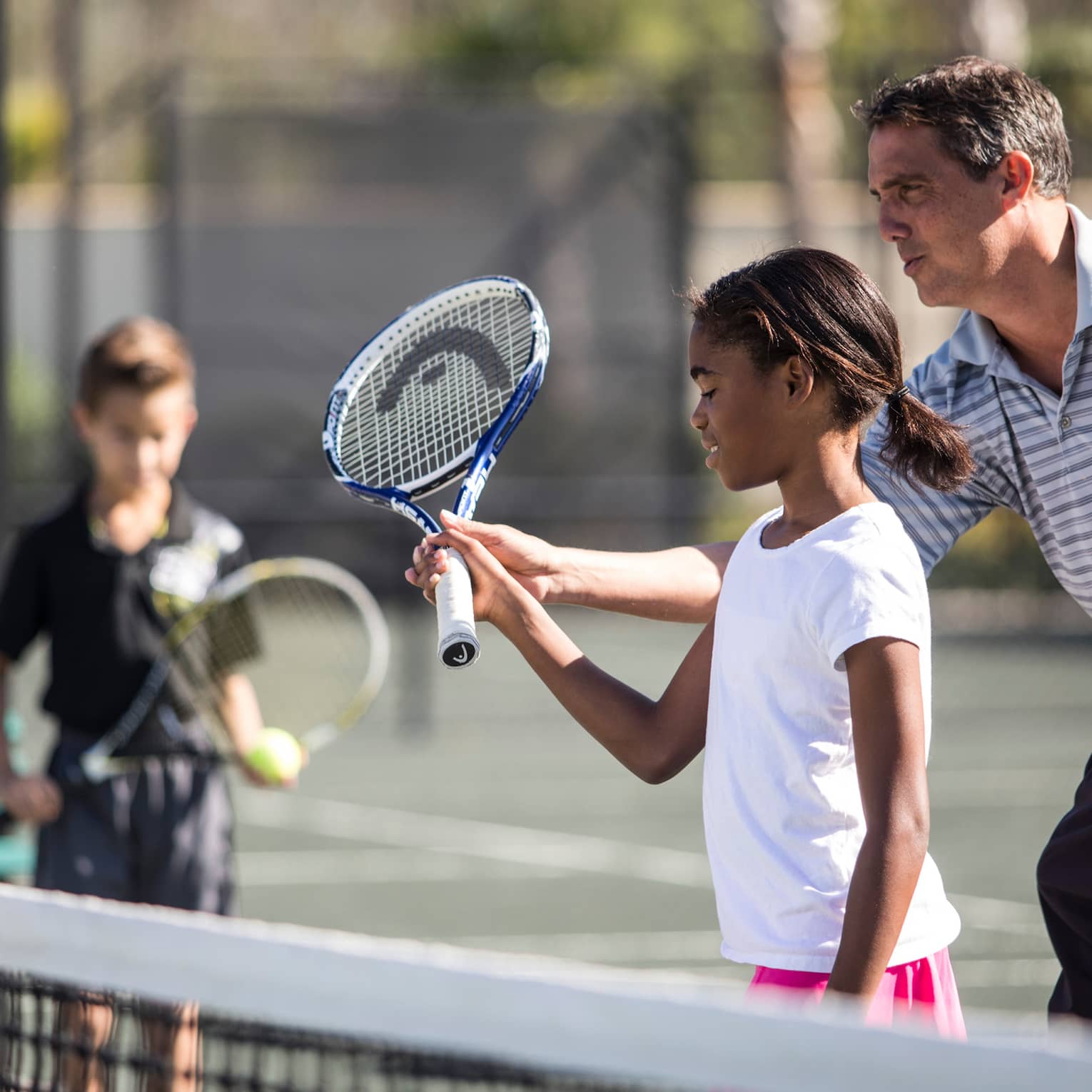Tennis coach helps young girl hold tennis racket by net as young boy watches