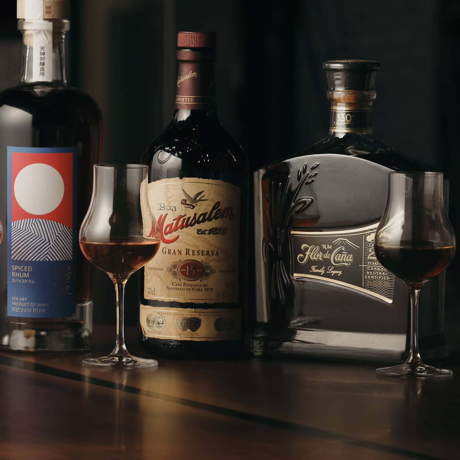 Dramatic dark lighting showcases three bottles of rum alongside three stemmed tulip-shaped glasses filled with dark rum.