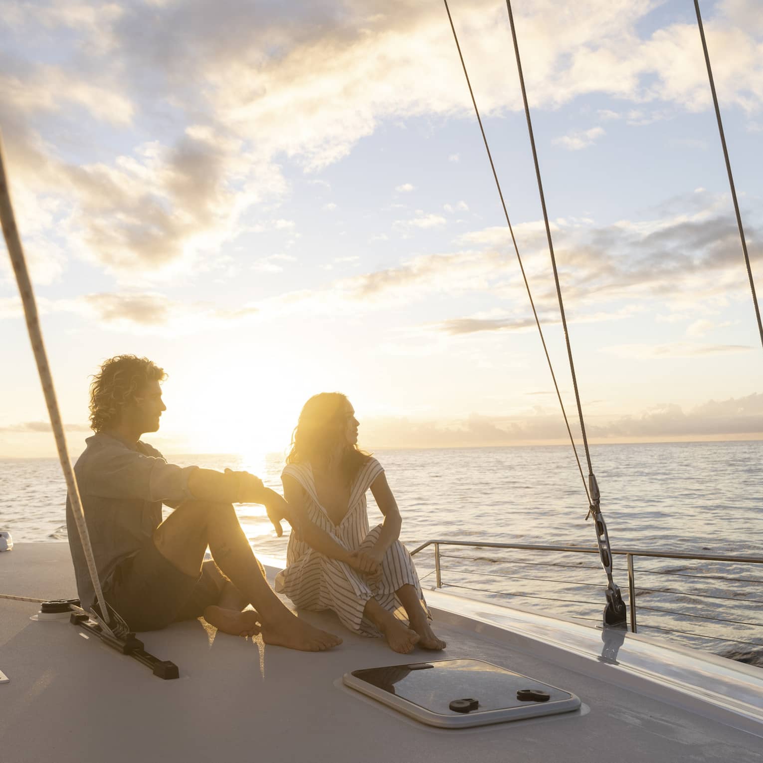 Couple in silhouette sitting barefoot on a boat deck framed by rigging ropes against a bright blue clouded sky at sunset.