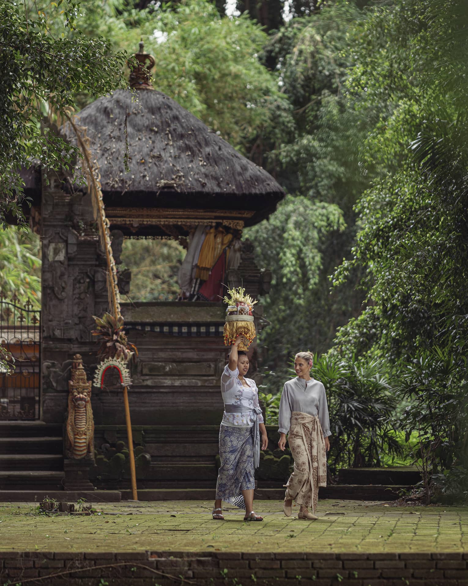 Two women wearing batik skirts walk by a temple in a lush tropical area as one of them carries a golden tray on her head.
