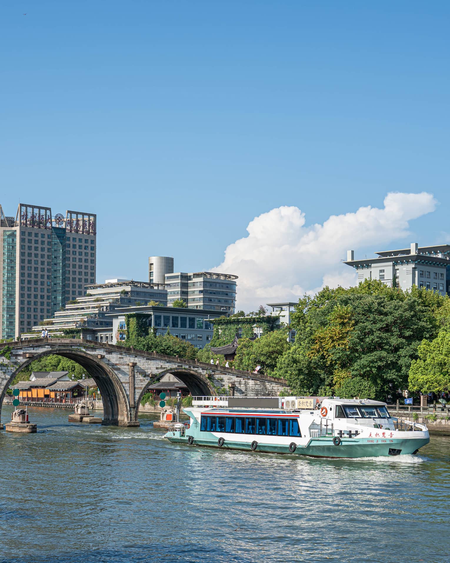 A tour boat passes under the centre arch of a historic stone bridge spanning a canal flanked by trees and modern buildings.