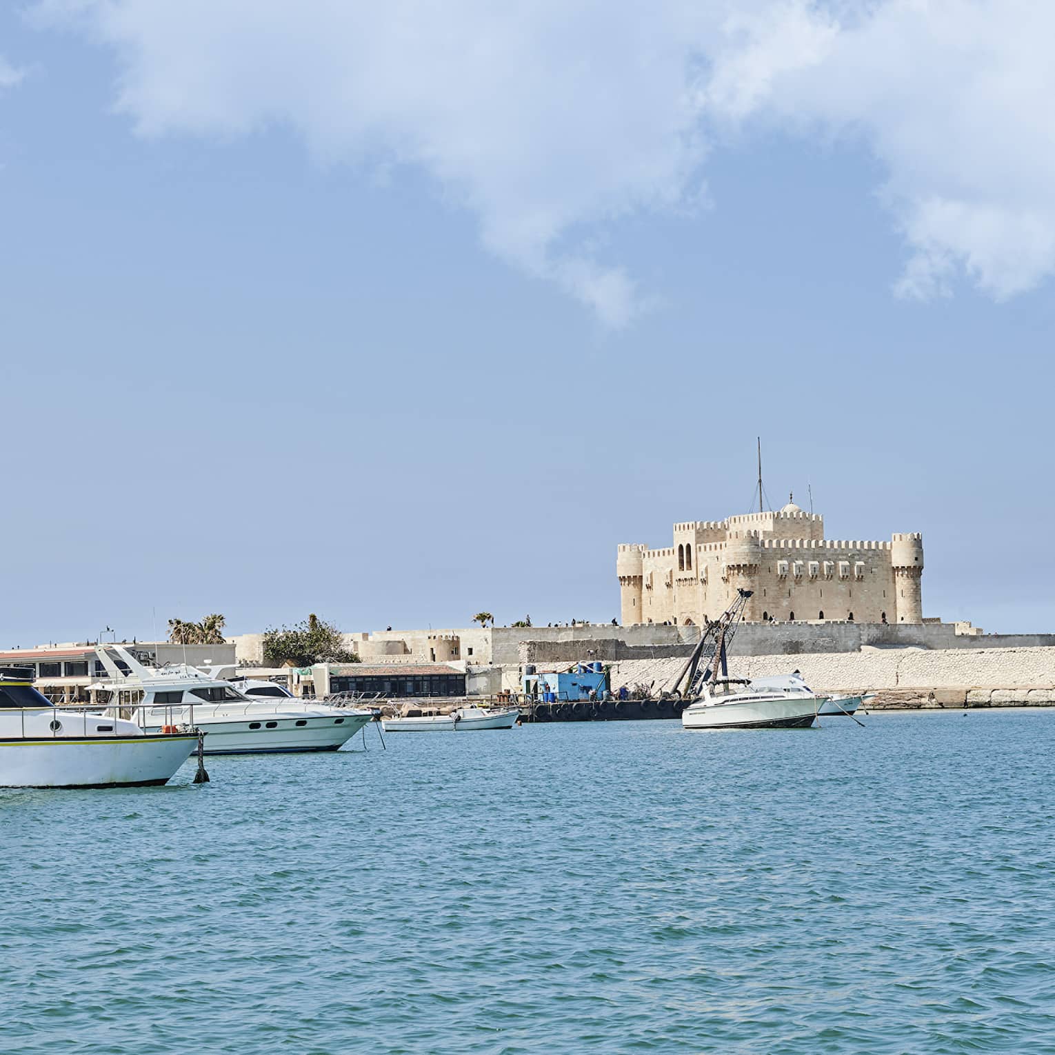A distant sand-coloured fortress perched on a quay along a shore dotted with small yachts and fishing boats over calm waters.