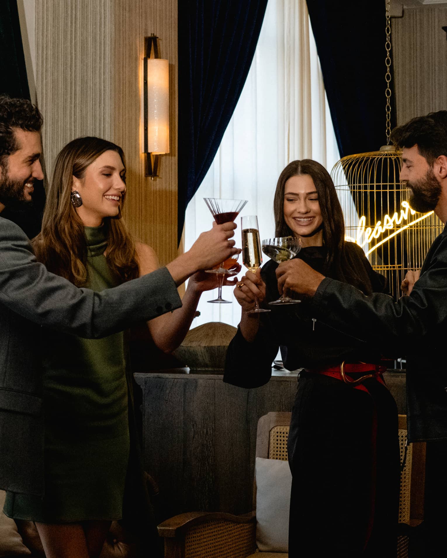 Four people raise their glasses in a toast in a cozy, softly lit lounge setting.