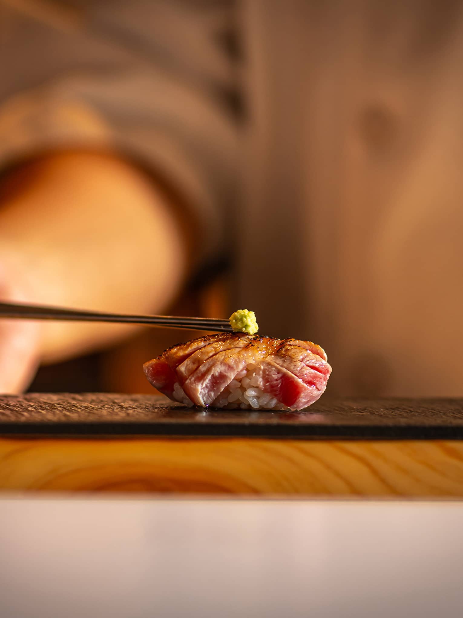 Seared tuna nigiri topped with a small dab of wasabi, placed on a black slate plate with a warm, blurred background