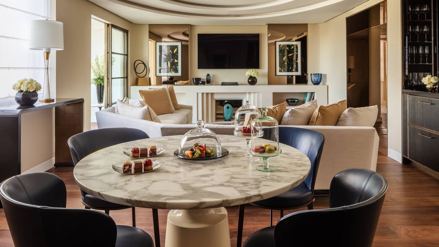 A suite with a round stone table with black chairs, a beige sofa, a television and large windows.