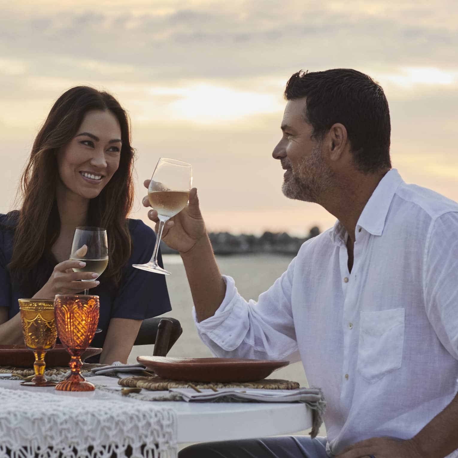 A smiling couple drinking wine against a sunset-streaked sky, at a beachfront dinner table set with amber-coloured goblets.