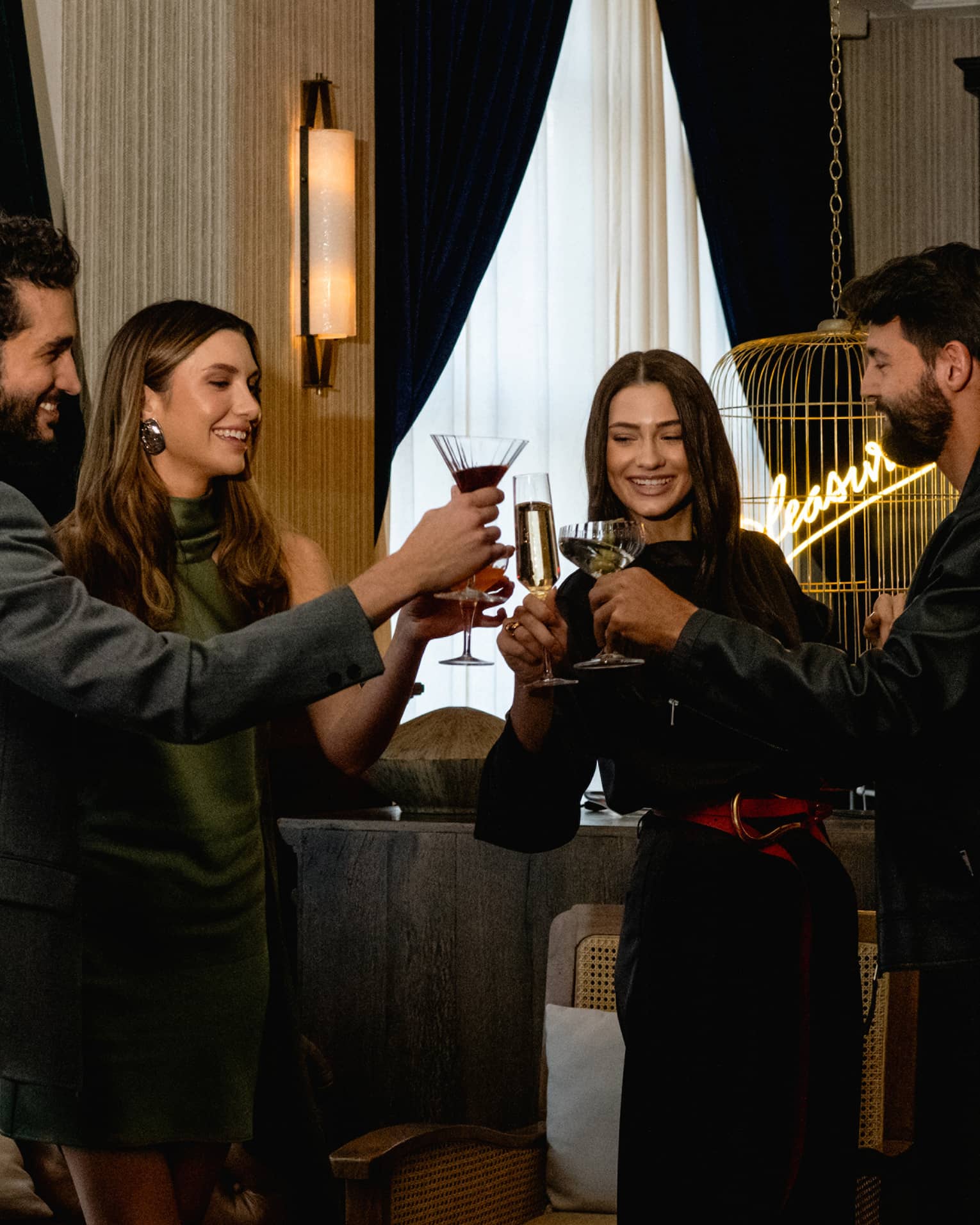 Four people raise their glasses in a toast in a cozy, softly lit lounge setting.