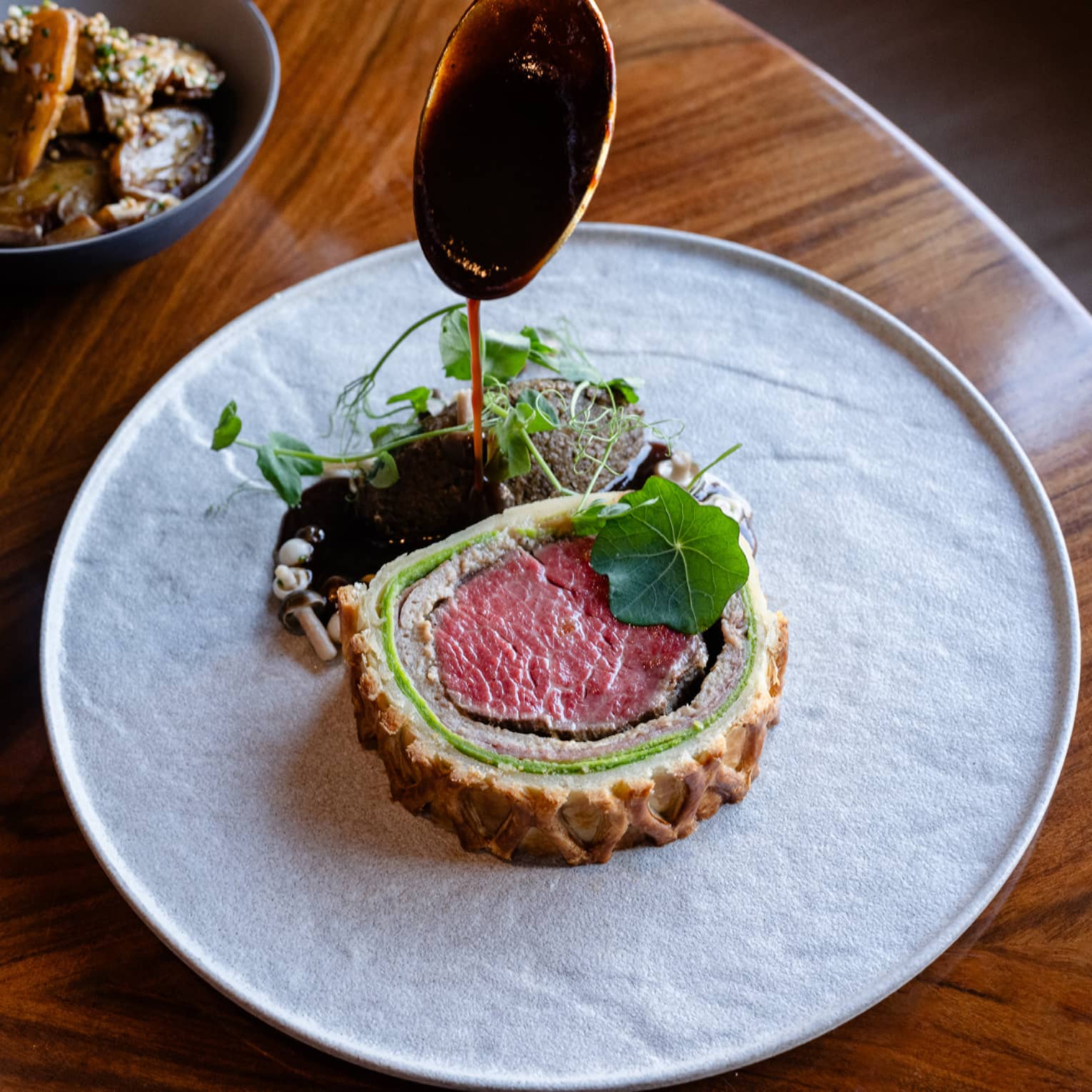 Slice of beef Wellington plated on a white dish with greens on the side plus a bowl of sliced vegetables and a bowl of mushrooms