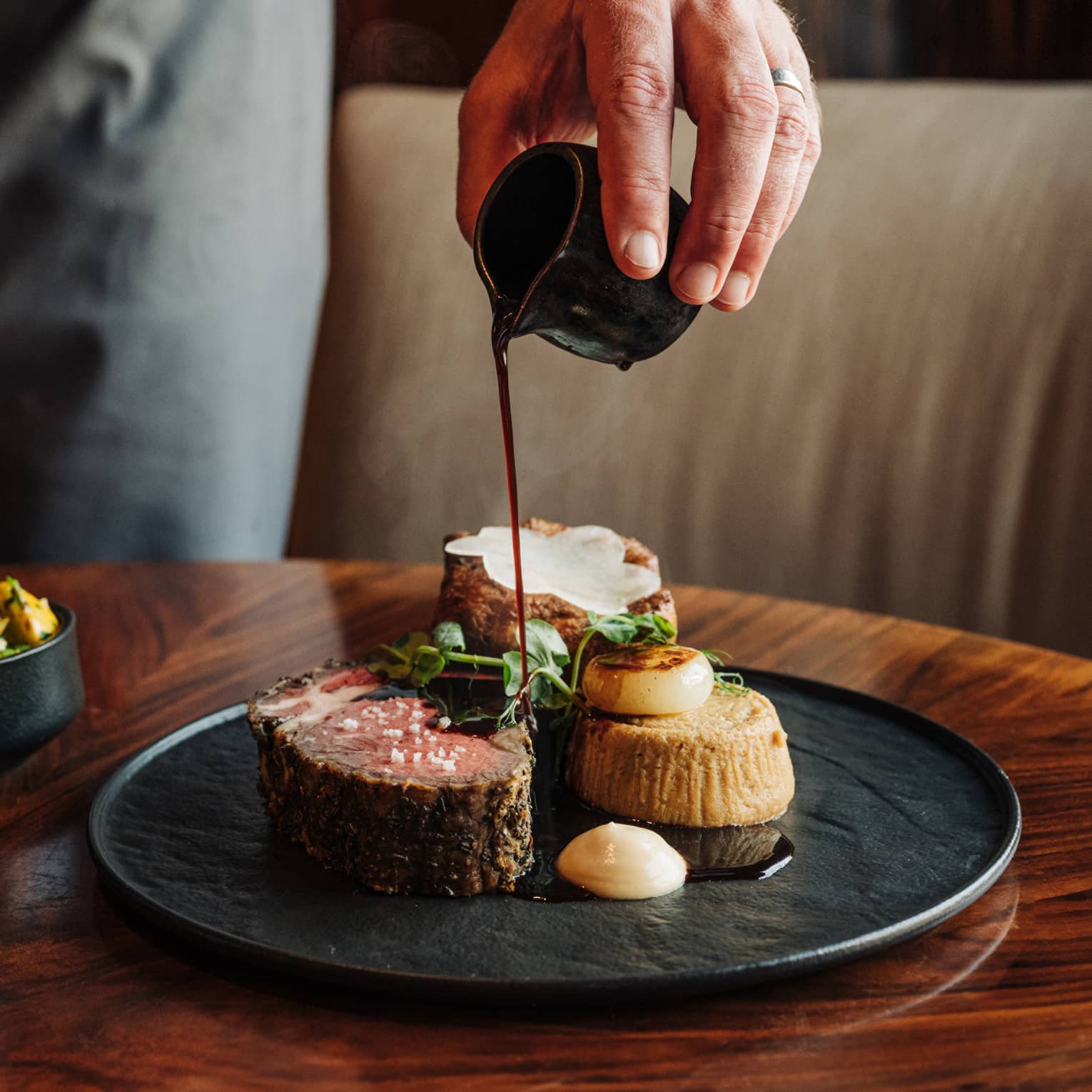 A waiter's hands pour sauce onto a slice of braised short rib served on a black plate next to two small black side bowls