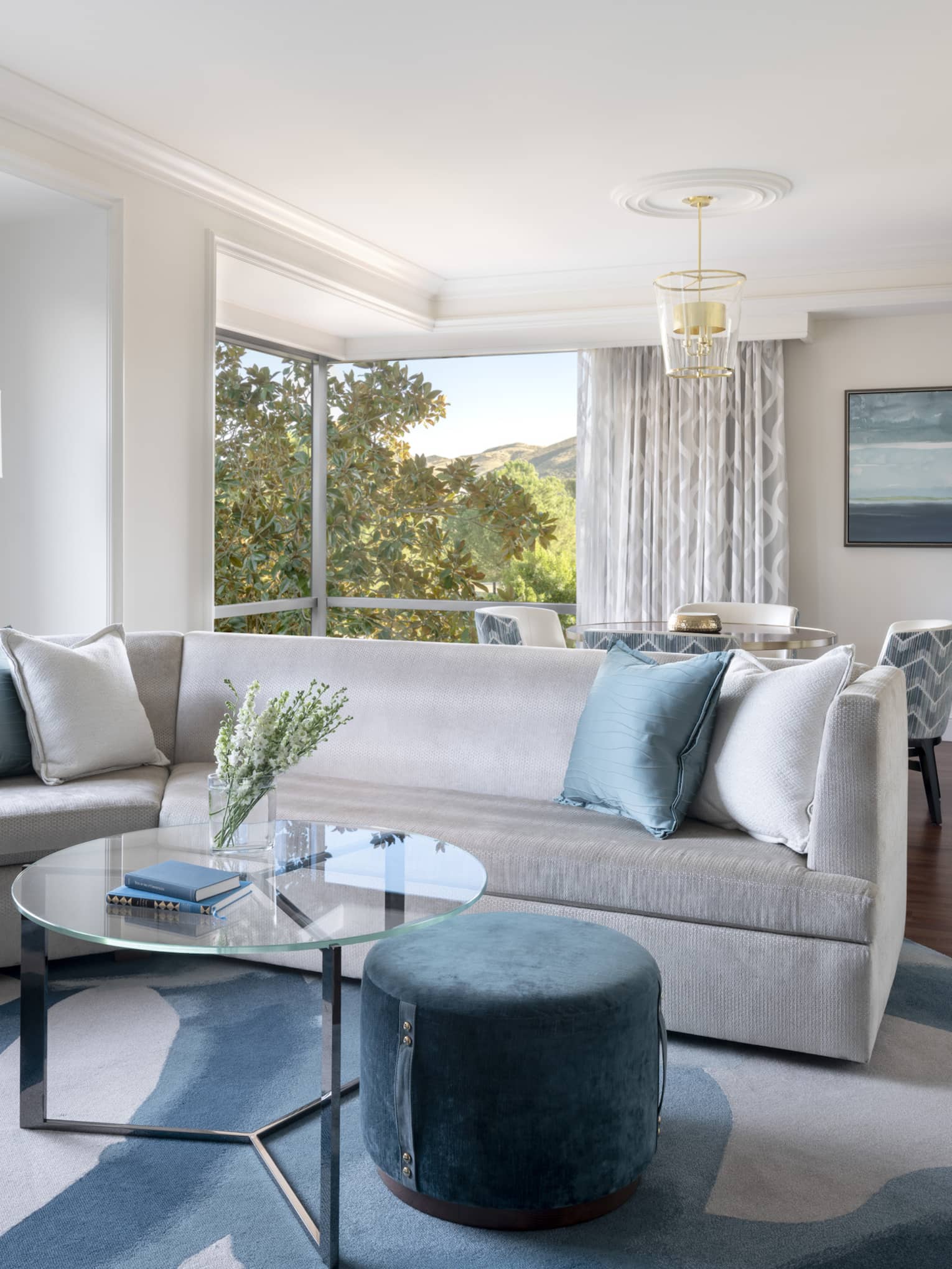Silver couch with blue pillows, a blue chair, glass coffee table, and a view of trees