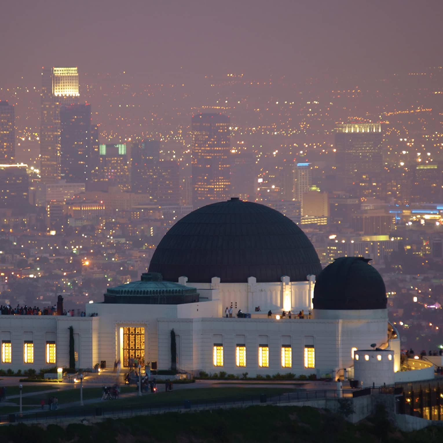 A large black and white observatory with three dome roofs, the city is behind it with many lights.